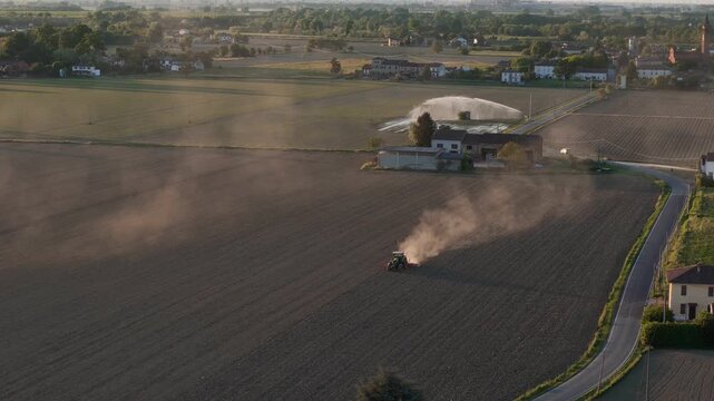 Agricultural activity in Emilia Romagna Pianura Padana Italy with tractor tillage generating dust across fields and sprinkler irrigation working near rural buildings drone parallax orbit aerial shot