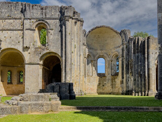 Fototapeta premium Visitors walk around the historic Abbaye de la Sauve-Majeur in Gironde, France. The ruins stand under a blue sky with clouds, revealing details of the architecture and scenery.