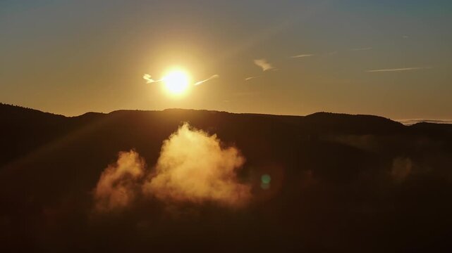Aerial drone shot of bright sun above dark mountain ridge, single isolated cloud glowing in warm golden light below ridge line, lens flare, golden sky