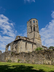 Visitors walk around the historic Abbaye de la Sauve-Majeur in Gironde, France. The ruins stand under a blue sky with clouds, revealing details of the architecture and scenery. © FreeProd