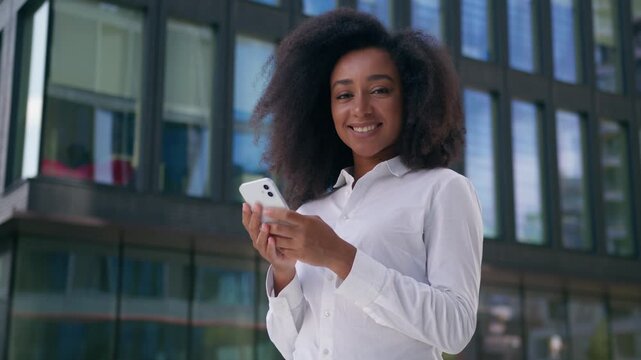 Cheerful African American business woman chatting online using mobile phone smartphone outdoors in city female smiling businesswoman texting message girl worker social media device looking at camera