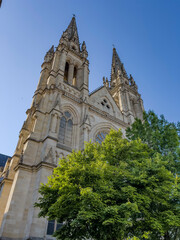 Saint-Louis Church rises above the trees in Quartier des Chartrons, Bordeaux. The church features tall spires and intricate details on its stone facade. The clear sky adds to its presence. © FreeProd