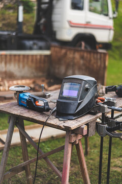 Assorted workshop tools including welding helmet and cutting discs placed on a wooden stool outdoors. Industrial DIY and metalworking equipment in natural light.