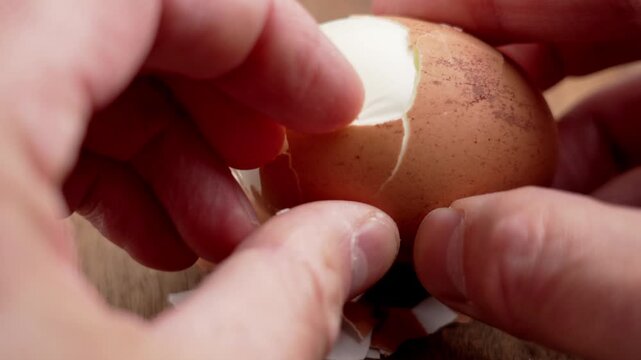 Closeup of human hands peeling a boiled chicken egg on wooden surface with eggshell fragments. Natural food preparation process