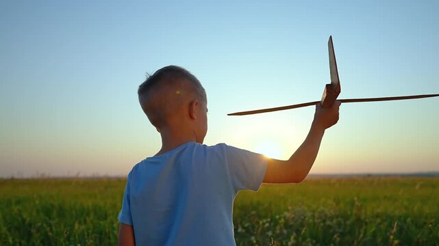 Boy holds wooden toy airplane overhead in field at sunset. Child plays with glider outdoors. Kid enjoys toy aircraft in countryside. Boy with wooden plane at golden hour. Child playing outside.