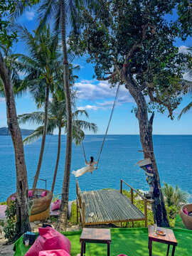 Young woman swinging above turquoise tropical sea surrounded by palm trees and relaxing island scenery