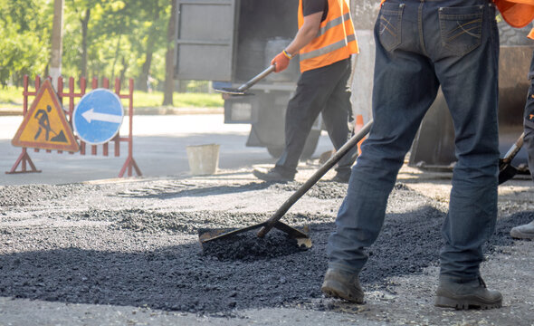 Road construction worker using a rake to level fresh asphalt with warning traffic signs in the background