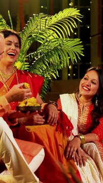 Holding laddus and smiling together, Indian family with young girl, senior mother in saree, father and husband in traditional wear celebrates Diwali indoors with diya, flower rangoli, and laughter