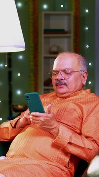 Surprising father with Diwali gift, Indian Asian young man surprising elderly dad with wrapped present while sitting together on decorated sofa indoors with warm lighting on Hindu festival celebration