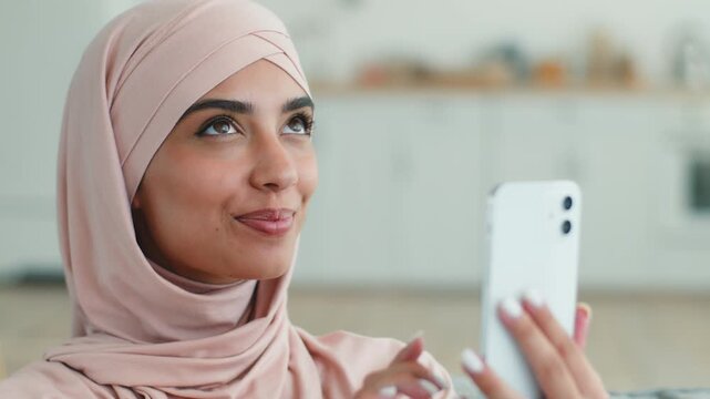 Smiling cheerful Arabian Muslim woman in hijab using mobile phone indoors female Islamic girl thinking choosing purchase shopping online browsing internet gadget addict social media influencer at home