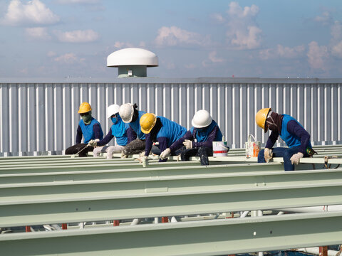 Team of workers in hard hats and safety vests works on building rooftop, painting metal roof framework. Industrial construction scene highlights teamwork, safety and construction project.