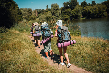 Obraz premium Three hikers with backpacks and sleeping mats walking along a lakeside trail on a sunny summer day