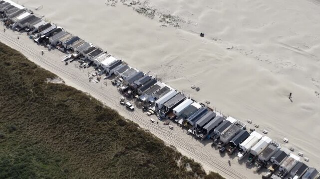 High aerial perspective moving along a long row of beach cabins situated between the sandy shoreline and grassy dunes, showing a unique coastal community and seaside living arrangement