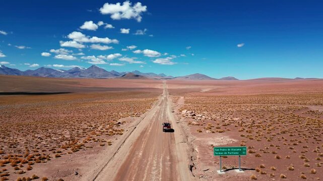 Tracking shot following a pickup truck driving along a straight desert dirt road past a road sign on vast arid altiplano with volcanic peaks on the horizon