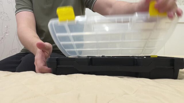 Hands opening plastic toolbox with yellow latches on bed, man arranging compartments and storing screws and fasteners, neutral wall background, soft indoor light, methodical DIY prep scene