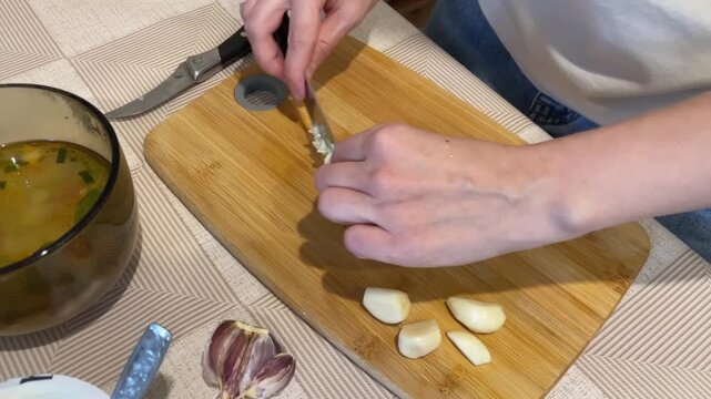 Hands mincing fresh garlic on cutting board, closeup of fingers chopping cloves beside bowl of soup, wooden board with peeled cloves, sharp knife, calm home kitchen atmosphere, savory aroma