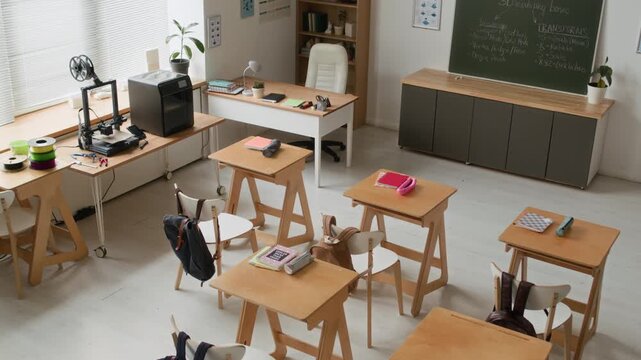 High angle tilt up no people shot of bright and functional modern classroom equipped with wooden desks for students, technology area with 3D printers, and greenboard with lesson notes