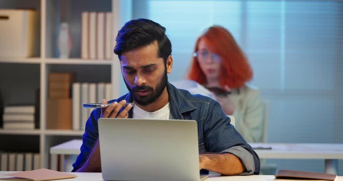 Diverse office workers speaking into smartphone interacting with virtual AI assistants while working on a laptop. Future technology, speech recognition, digital automation, smart corporate office.