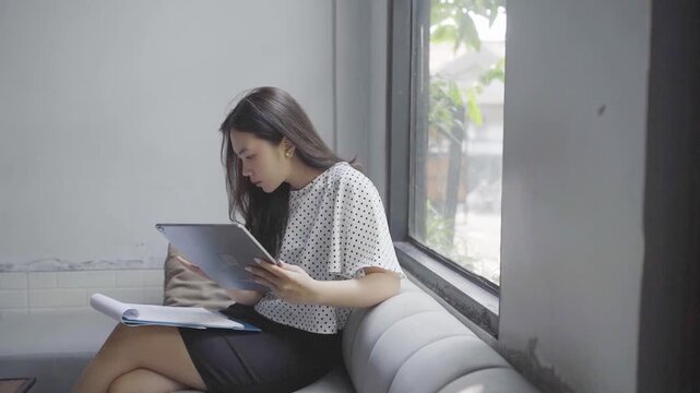 Asian woman reading tablet by window, soft natural light, urban view outside, flipping through printed notes and annotating pages,
