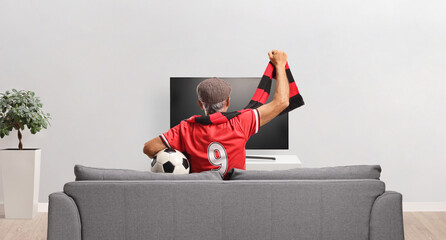 Obraz premium Rear view shot of a senior sports fan in a red jersey cheering with a scarf on a sofa