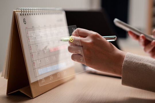 Time management and workflow scheduling, digital calendar overlay. Woman marking appointment dates on a desk calendar while holding a smartphone, planning deadlines and tracking daily tasks.