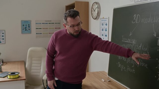 Bearded male teacher pointing to chalkboard with notes on 3D modeling basics while giving lecture to student in school