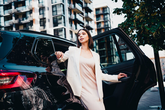 Businesswoman stepping out of luxury SUV in urban setting, stylish executive wearing blazer and dress, confident modern professional in city environment near apartment buildings