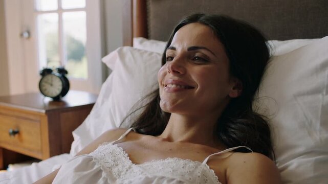 Smiling woman reclining in bed, seeing dawn light, looking right in white camisole and alarm clock