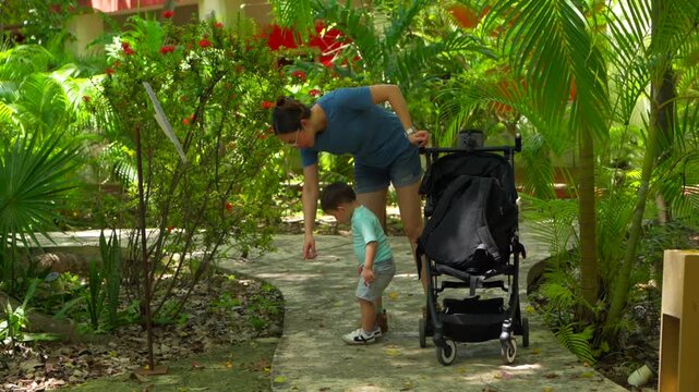 A loving Latin mother and her toddler explore a vibrant tropical garden at a museum. The mom points to objects on the ground and then to the tall trees, fostering a sense of wonder and discovery.