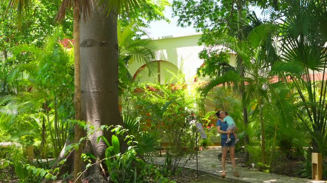 A young mother carries her baby through the lush museum garden, observing a tall ceiba tree. This scene captures a moment of family exploration and cultural discovery.