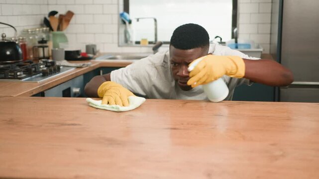 Young Man Happily Cleans Kitchen Countertop with Spray and Cloth, Achieving a Spotless Finish