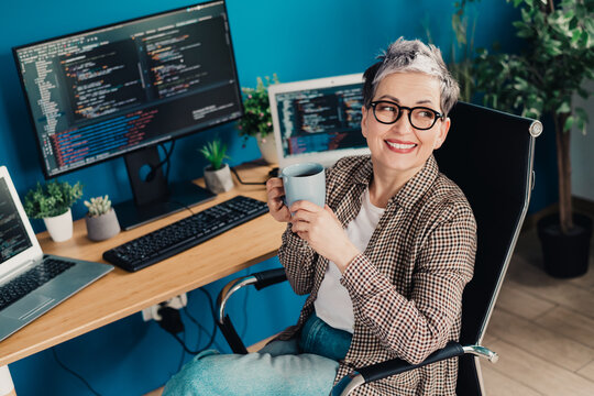 Smiling middle-aged female programmer coding in a home office with dual monitors and laptop while holding a coffee mug, casual remote work setup with plants and ergonomic chair