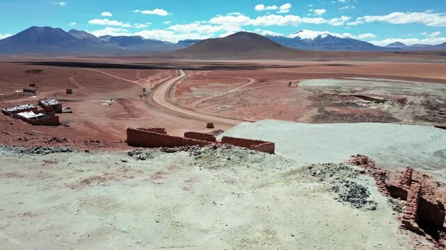 Dolly shot gliding over ancient atacame&ntilde;o stone corral ruins on red desert plateau with winding dirt road and dramatic snow-capped volcanic peaks under blue sky. Putana, Chile