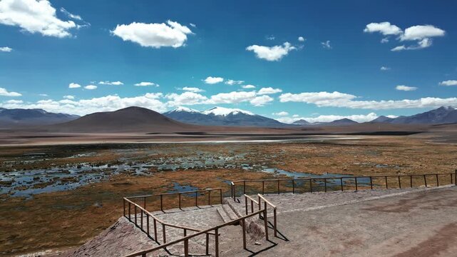 Orbit over rustic lookout platform overlooking vast altiplano bofedal wetland with lagoon and snow-capped volcanic peaks stretching to the horizon. Putana, Chile
