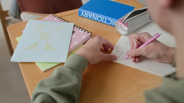 High angle over the shoulder shot of unreconizable boy writing in his notebook while taking notes at desk during school class