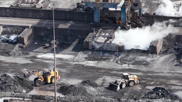 High angle view of two front end loaders moving piles of coal at an industrial plant, with thick white smoke rising from a quenching station in a vast steel mill yard during production