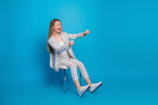 Young businesswoman in a checkered suit sitting on a chair against a blue background pretending to drive while laughing and gesturing confidently