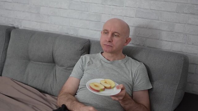 Bald man relaxing on sofa and eating fresh apple slices at home Full HD video