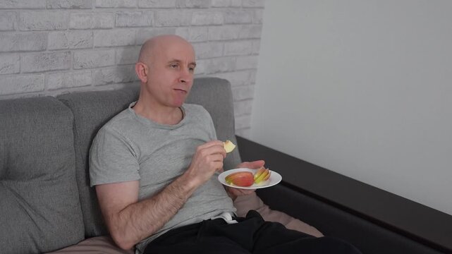 Bald man relaxing on sofa and eating fresh apple slices at video HD Full home