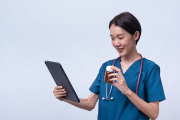 Fototapeta premium Smiling Asian female doctor in scrubs holding a tablet and medicine bottle. Online pharmacy and telehealth prescription concept isolated on white background.