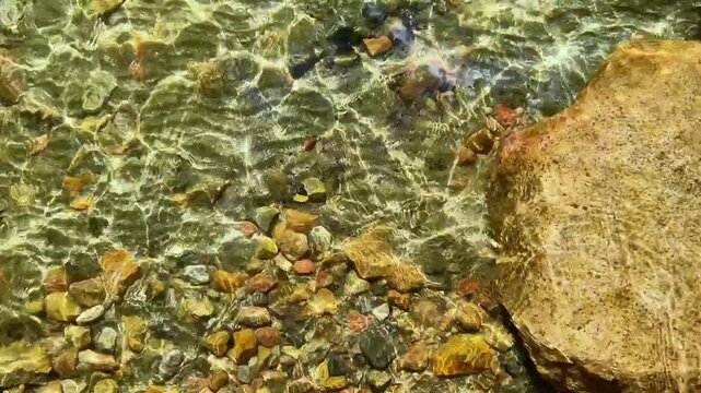 Clear water surface revealing colorful stones and textured rock beneath, illuminated by bright natural light.