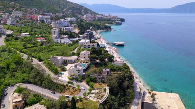 Aerial video over the rugged coastal edge of Vlora, Albania, showcasing the promenade, winding hillside roads, coastal hotels, and the blue Ionian Sea. 