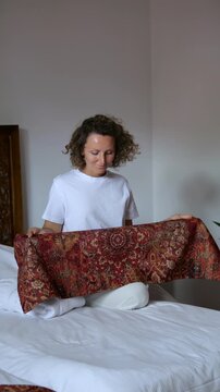 Smiling woman with curly hair tidying up the bedroom, sitting on a white bed and carefully folding a beautiful ornamental bed runner as part of her morning household chores and routine