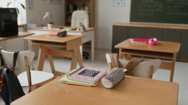 Close up no people shot of pink notebook and gray pencil case on wooden desk of student, and other desks and black 3D printer in modern classroom