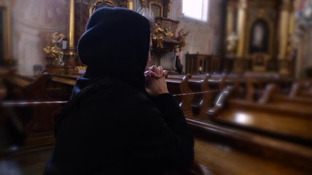 Young woman wearing a black hood praying with her hands clasped on a wooden pew inside a beautiful and ancient christian church, seeking solace and faith in a moment of quiet contemplation