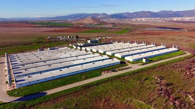 Drone view of livestock farm buildings in valley, Bulgaria. White and silver roofed barns, green perimeter grass, red brown plowed fields and distant city against mountain range under blue sky.