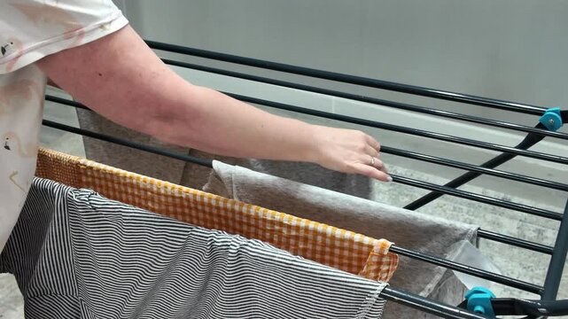 An unrecognizable woman doing laundry, hanging wet clothes on a folding drying rack indoors. Concept of household chores and routine