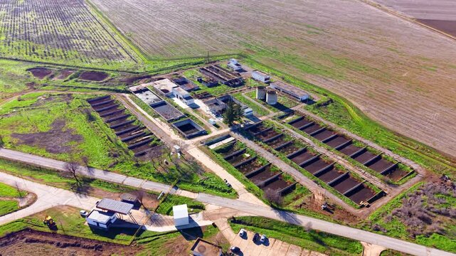 Drone view of wastewater treatment plant at farm animal complex. Oblique aerial of rectangular settling tanks, cylindrical digesters, white utility buildings and dirt roads amid green fields.