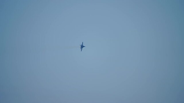 Distant silhouette of a twin-engine military fighter jet maneuvering in a hazy grey sky with faint vapor trace at the wingtip
