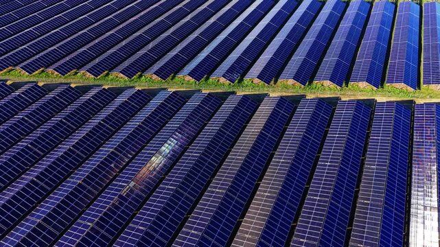 Long photovoltaic rows oriented diagonally with hard midday shadows and a lens flare accent point. Drone view of solar farm panels with strong diagonal shadows
